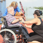 Man in wheelchair getting assistance to shoot a bow and arrow.