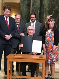 Governor Evers holding up signed bill sitting at table surrounded by people standing